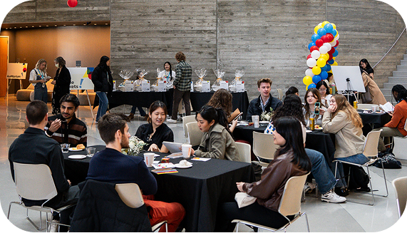 An open atrium space in the conference venue is filled with multiple tables covered with tablecloths, each seating several students. Attendees are engaged in conversations, primarily in pairs with the students next to them. In the background, a balloon arch stands near a table where student volunteers are stationed behind an array of gift baskets, interacting with attendees