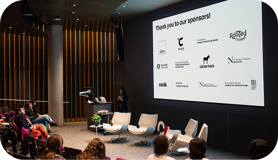 The previous conference director for Interventions stands on a stage in a dimly lit auditorium in the ISEC building, which is the venue for the event, presenting to a crowd. Behind her, a large slide displays the words “Thank you to our sponsors!” along with several sponsor logos.