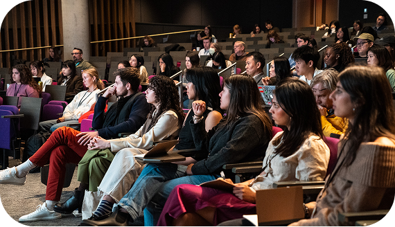 An auditorium filled mainly with students, all looking ahead at the stage. Some are taking notes, while others listen attentively, appearing deeply focused on the presentation.