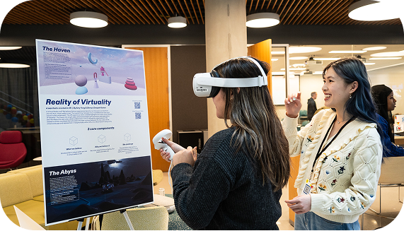 A Northeastern student presenter smiles while showcasing a poster titled Reality of Virtuality at an event. To her left, another student, wearing a VR headset and holding a controller, interacts with the virtual reality experience while facing the poster.